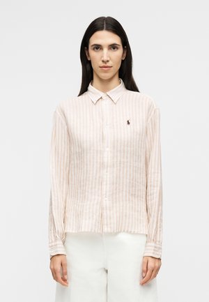 Γυναίκα με μακριά σκοτεινά μαλλιά φοράει πουκάμισο με κάθετες ρίγες σε μπεζ και άσπρο χρώμα και άσπρο παντελόνι, στέκεται μπροστά σε απλό φόντο.