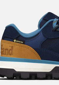 Zapato de senderismo azul marino con etiqueta GORE-TEX, material de gamuza y malla, acentos en azul claro, sección del talón marrón y suela de goma con patrón.