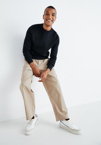 Mørkeblå strikkegenser, beige vide bukser og hvite sneakers med grå detaljer. Modellen sitter med hendene på knærne.