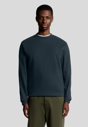 Donkerblauwe sweatshirt met ronde hals, lange mouwen en een subtiele logodetail op de borst. G gedragen met olijfgroene broek.