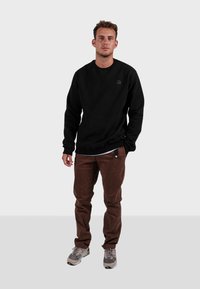 Černá mikina s kulatým výstřihem a malým logem, doplněná o hnědé kalhoty a šedé tenisky, které kombinují různé textury a barvy.