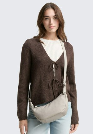 Brauner strukturierter Pullover mit V-Ausschnitt und vorn gebundenen Akzenten, über einem weißen gerippten Top getragen. Das Modell hält eine hellgraue Schultertasche.