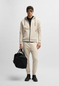 Sweatshirt beige à fermeture éclair avec cordons, associé à un pantalon assorti et des baskets noires. Porte un sac de sport noir. Le look est athlétique et épuré.