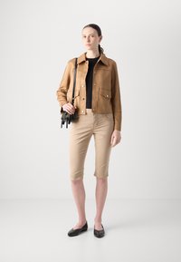 Brun semsket skinnjakke med trykknapper, svart tettsittende topp, beige knelange shorts og svarte flate sko. Modellen holder en liten svart skulderveske.