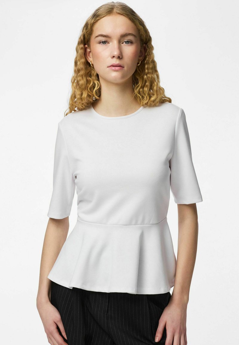 Jeune femme aux cheveux blonds bouclés portant de petites boucles d'oreilles créoles, un haut blanc à manches courtes avec basque, et un pantalon noir à fines rayures sur fond blanc.