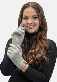 Grijze gebreide handschoenen met een geribbeld oppervlak en een leren accentlabel, gedragen door een persoon met lang, golvend haar, tegen een effen achtergrond.