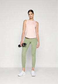 Light pink sleeveless athletic top paired with high-waisted olive green leggings. Model holds a black water bottle and wears white sneakers.