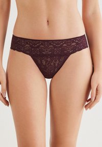 Kobieta w ciemnofioletowej koronkowej bieliźnie bikini o niskim stanie, eksponująca środkową część ciała i górne uda na jednolitym tle.