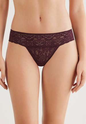 Vrouw in een donkerpaarse kanten lage taille bikini onderbroek, die de middensection en bovenbenen toont tegen een effen achtergrond.