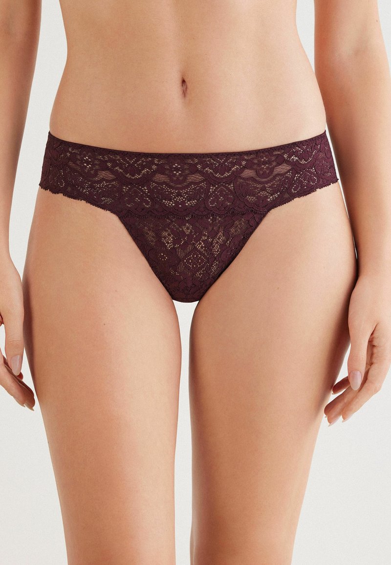Kobieta w ciemnofioletowej koronkowej bieliźnie bikini o niskim stanie, eksponująca środkową część ciała i górne uda na jednolitym tle.