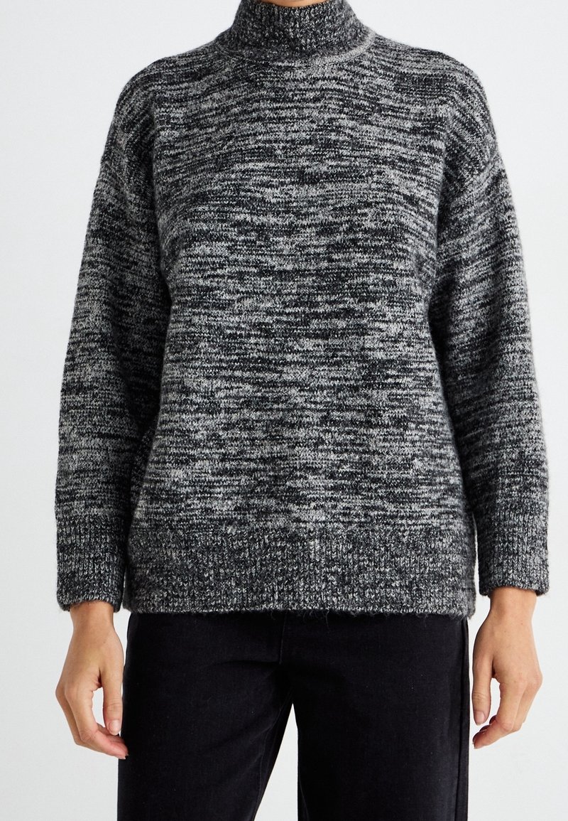 Personne portant un pull à col roulé tricoté gris et noir à manches longues, associé à un pantalon noir, sur un fond uni.