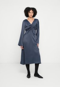 Rochie satinată bleumarin închis cu decolteu în V, mâneci lungi și detaliu frontale adunat. Țesătura are o textură netedă și lucioasă.