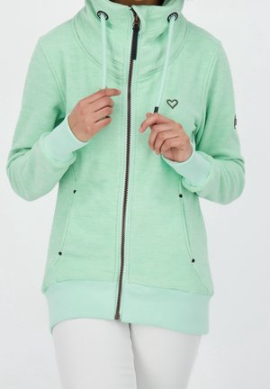 Personne portant un sweat à capuche zippé vert menthe avec un emblème en forme de cœur, tenant les cordons de la capuche près de la poitrine.