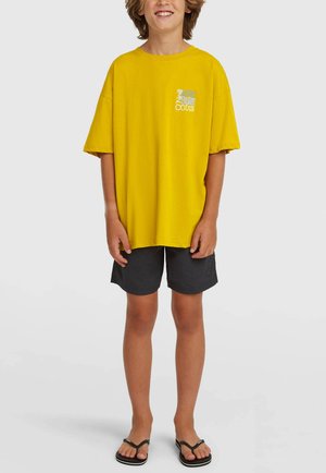 Kollane puuvillane T-särk lõdva lõikega, lühikeste varrukatega ja väikese graafilise detailiga rinnal. Sobitatud tumedahallide lühikeste pükstega ja plätudega.