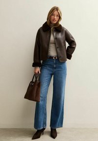 Bruine faux leren jack met een bontkraag, beige top, wijde blauwe jeans en een bruine textuurhandtas. Donkere puntige hakken maken de outfit compleet.
