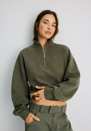 Femme aux longs cheveux bruns portant un sweat-shirt court zippé vert olive et un pantalon assorti, debout devant un fond uni.