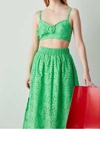 Haut court vert avec un accent circulaire et jupe assortie présentant des motifs à œillets et une taille froncée. Sac fourre-tout rouge tenu à la main.