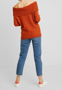 Pull orange rouille à épaule dénudée avec un col côtelé, associé à un jean court bleu clair et des baskets blanches avec des accents orange.
