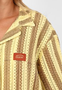 Chemise à rayures jaunes et brunes avec un design texturé, présentant des motifs découpés, un col et une petite étiquette rouge OXBOW.