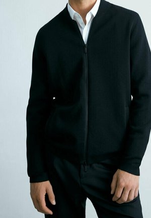 Mężczyzna w czarnej kurtce zapinanej na zamek na białą koszulę z kołnierzykiem, stojący z rękami swobodnie opuszczonymi wzdłuż ciała na jednolitym tle.