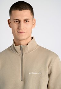 Sudadera beige con cremallera y cuello alto, con una cremallera plateada y texto de logo blanco en el lado izquierdo. Tejido suave y texturizado.