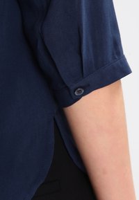 Blusa blu navy con una consistenza morbida; presenta maniche arrotolate e un dettaglio con bottoni, che mette in risalto un design minimalista e una vestibilità su misura.