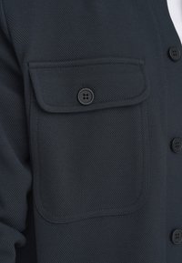 Mörkblå knapplappad skjorta med en framträdande bröstficka, texturerad tyg och kontrasterande svarta knappar.