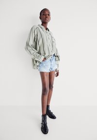 Camisa de rayas verdes de gran tamaño con cierre de botones, combinada con pantalones cortos de denim desgastados. Botas de tobillo con hebilla negras. Fondo simple.