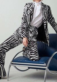 Personne portant un costume rayé zèbre noir et blanc et une blouse blanche à volants, agenouillée sur une chaise moderne en cuir bleu avec un cadre chromé.