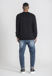 Sudadera negra de manga larga, con puños y dobladillo acanalados. Combinada con jeans azules desgastados y zapatillas negras con patrón. Fondo blanco.