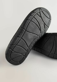 Semelles de chaussures en caoutchouc noir avec une surface texturée, présentant un motif de bosses et de rainures pour l'adhérence et la traction.