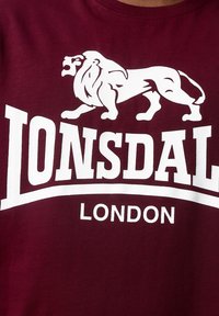 Tricou din bumbac închis la culoare, cu un grafic mare alb al unui leu deasupra textului "LONSDALE" și "LONDON", având o textură netedă și guler rotund.