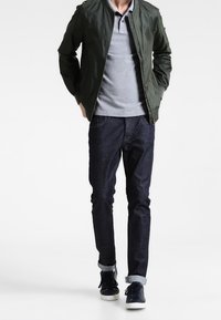 Zielona kurtka typu bomber na szarej koszulce polo, do tego ciemne dżinsy i czarne trampki z białymi podeszwami. Klasyczny, casualowy strój.