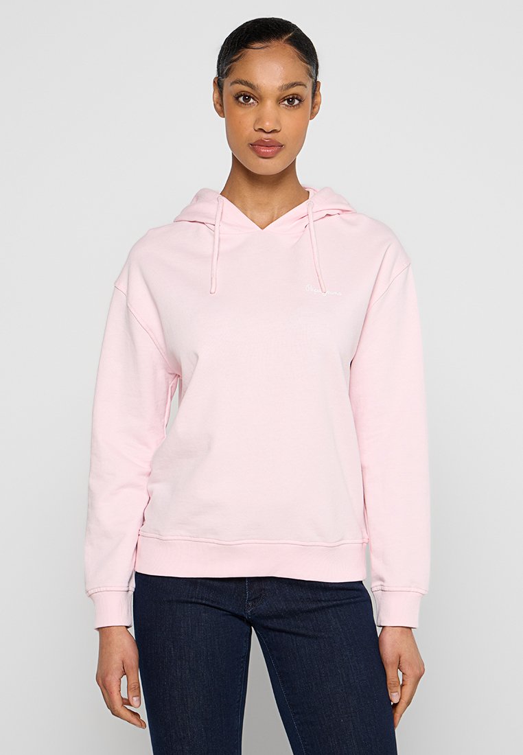 Junge Frau in einem hellrosa Kapuzenpullover mit Kordelzug und dunkelben Jeans, steht vor einem schlichten, hellgrauen Hintergrund.