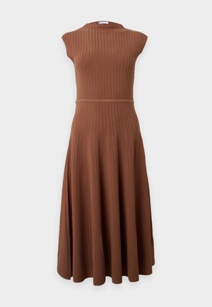 Femme aux longs cheveux foncés portant une robe midi marron sans manches côtelée et des chaussures plates noires, debout devant un fond clair uni.