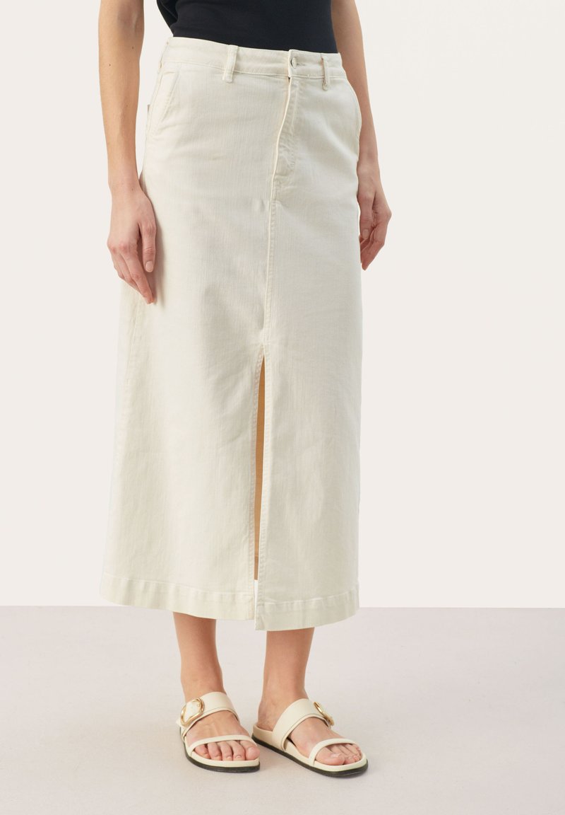 Jupe en denim beige clair avec une fente sur le devant, taille haute et fermeture à boutons. Associée à des sandales plates de couleur claire.