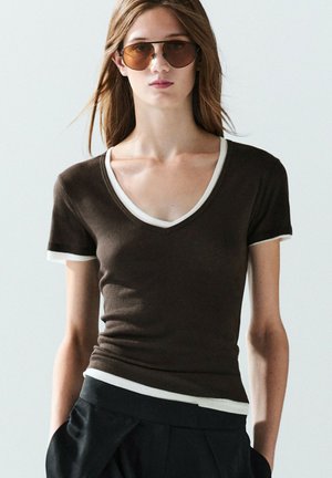 Femme portant un t-shirt marron col V à manches courtes avec un bord blanc et de grandes lunettes de soleil marron, debout devant un fond clair uni.