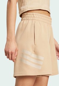 Pantalones cortos deportivos beige con una cinturilla elástica, que presentan tres acentos en forma de franjas horizontales de un material brillante en el muslo izquierdo.