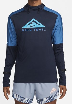 Mann trägt navyblaue und blaue Nike Trail Kapuzenpullover mit langen Ärmeln und hellblaue, gemusterte Nike Trail Laufshorts.