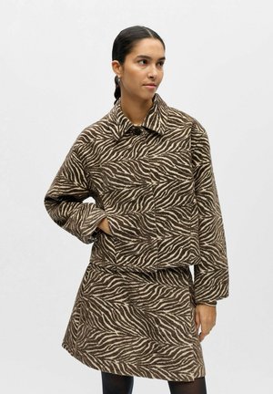 Femme portant une veste rayée zèbre marron et beige et une jupe assortie, debout avec une main dans la poche, regardant sur le côté.