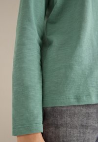 Vaalea vihreä pitkähihainen paita, jossa on sileä rakenne, pyöreä helma ja yksityiskohtainen ompelu. Pukeutunut tummanharmaan kankaan päälle.