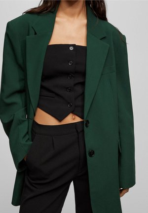 Femme portant un blazer oversize vert foncé sur un crop top noir sans bretelles et boutonné, ainsi qu'un pantalon noir taille haute, les mains dans les poches.