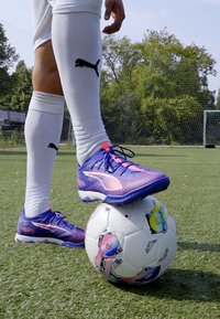 Puma nogometne kopačke s dizajnom u ljubičastoj i ružičastoj gradaciji, s crnim vezicama, odmaraju na uzorčanoj nogometnoj lopti na umjetnoj travnatoj površini.