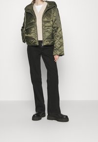 Quiltet olivengrøn pufferjakke med hætte, med to forlommer, båret over en lys beige sweater og sorte bukser, parret med chunky sorte støvler.