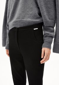 Schwarze Hose mit einem Knopfverschluss vorne, weicher Textur und Seitentaschen, kombiniert mit einem lockeren, grauen Strickpullover.