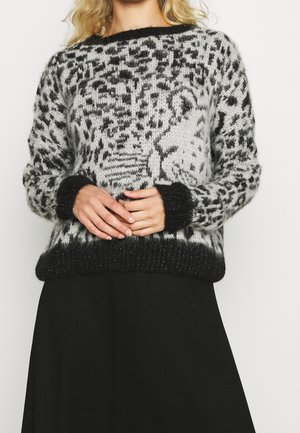 Femme portant un pull pelucheux gris et noir avec un motif de visage de léopard, associé à une jupe noire, mains jointes à la taille.