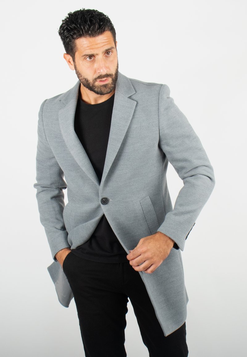 Blazer ajusté gris avec une fermeture à un bouton, des revers à cran et deux poches avant. Porté sur une chemise noire et associé à un pantalon noir.