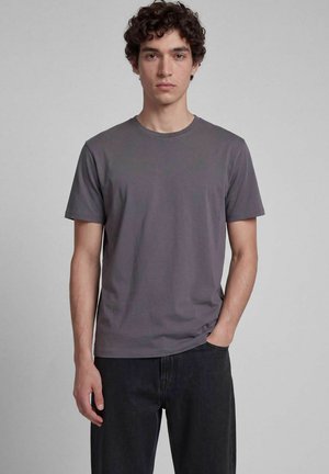 Jeune homme aux cheveux bouclés portant un t-shirt gris uni à manches courtes et un jean noir, debout devant un fond neutre.