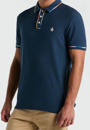 Homme portant un polo texturé bleu marine avec un liseré arc-en-ciel au col et aux manches, et un pantalon beige, debout devant un fond uni.