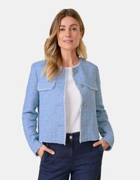 Hellblaue Boucle-Jacke mit fransigen Kanten, zwei Fronttaschen und silbernen Knöpfen. Getragen über einem weißen Oberteil und kombiniert mit dunkelblauen Jeans.